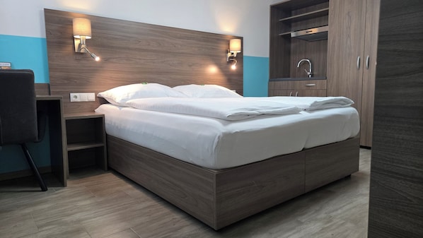 A modern hotel room with a wooden headboard, a bed with white linens, a nightstand, a desk, and a wardrobe with a mirror. - Hotel König (Vienna)