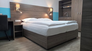 Una camera d'albergo moderna con una testiera in legno, un letto con lenzuola bianche, un comodino, una scrivania e un armadio con uno specchio.