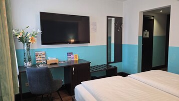Una camera d'albergo con un letto, una scrivania con una sedia, una televisione, uno specchio e un vaso con fiori.