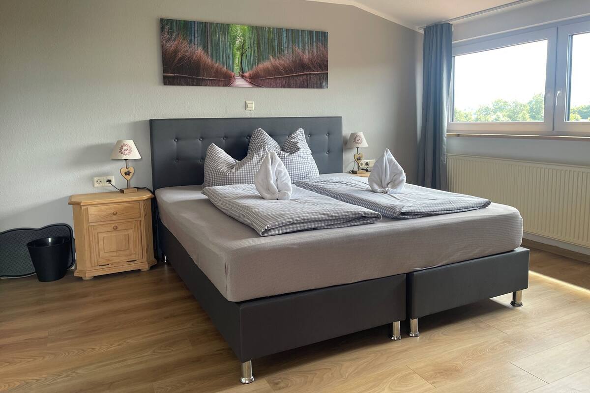 Une chambre moderne avec un grand lit, deux tables de chevet, une commode, une fenêtre avec des rideaux et une œuvre d’art accrochée au mur.