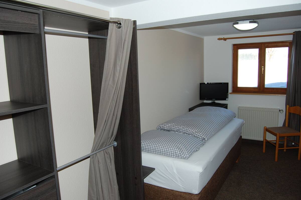 Une chambre avec un lit simple, une télévision, une chaise et une fenêtre avec des rideaux.