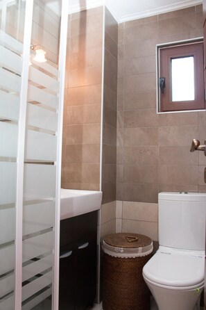 A bathroom with a toilet, a sink, a trash bin, and a shower area. - Villa Vlatos (Kissamos)