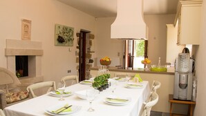 A dining area with a table set for four, a fireplace, stone walls, and a kitchen area with a water dispenser. - Villa Vlatos (Kissamos)