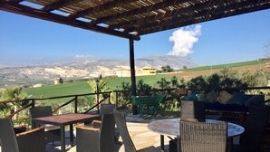 Eine Terrasse im Freien mit Korbmöbeln und Blick auf ein grünes Feld und ferne Berge.