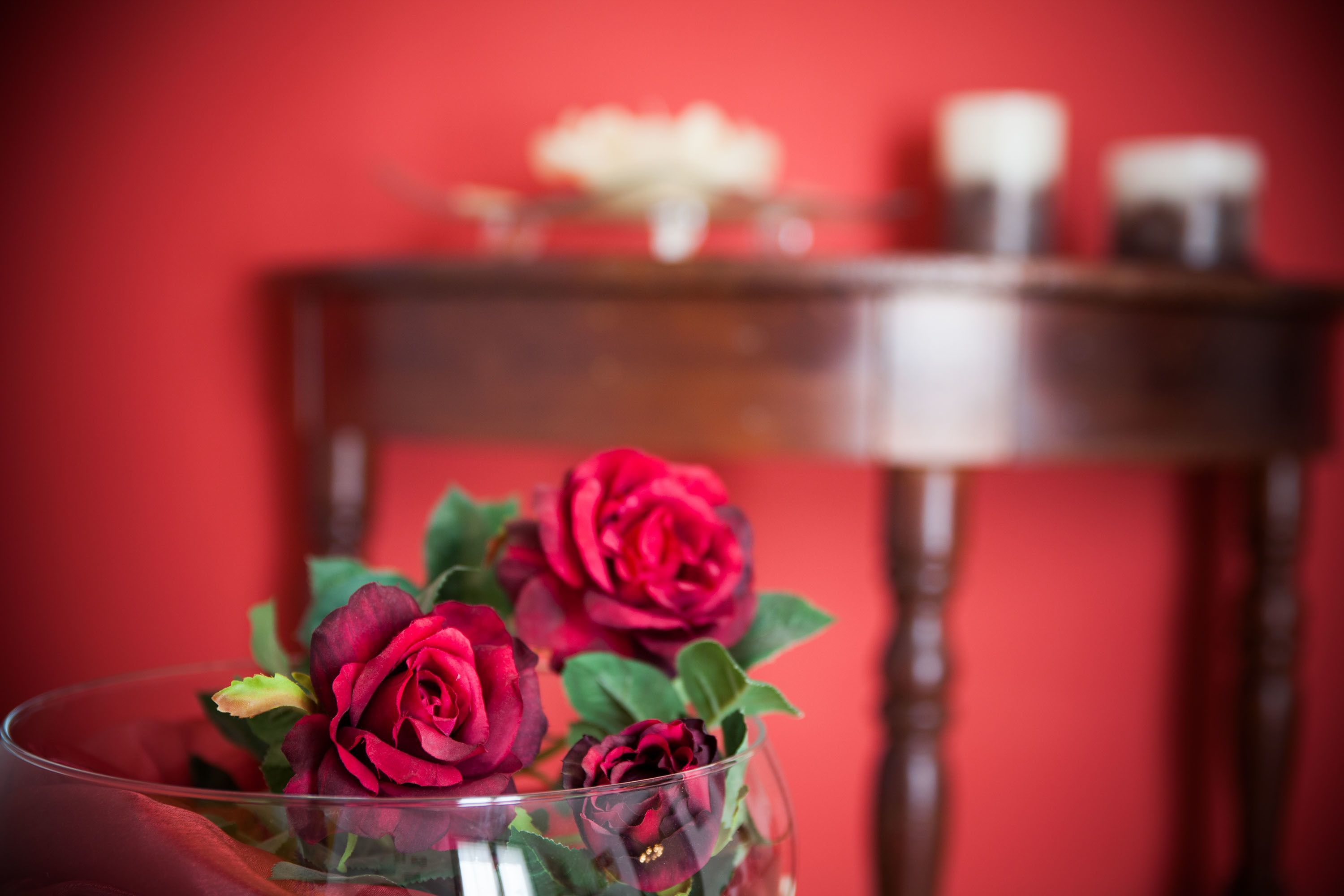 A glass vase with red roses on a table against a red wall.