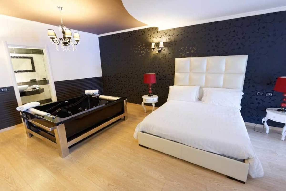 A modern bedroom with a large bed, a separate bathtub, a mirror, and a chandelier.