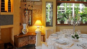 Interior de restaurante con pared de piedra, una mesa dispuesta para comer, una lámpara y una ventana con plantas.