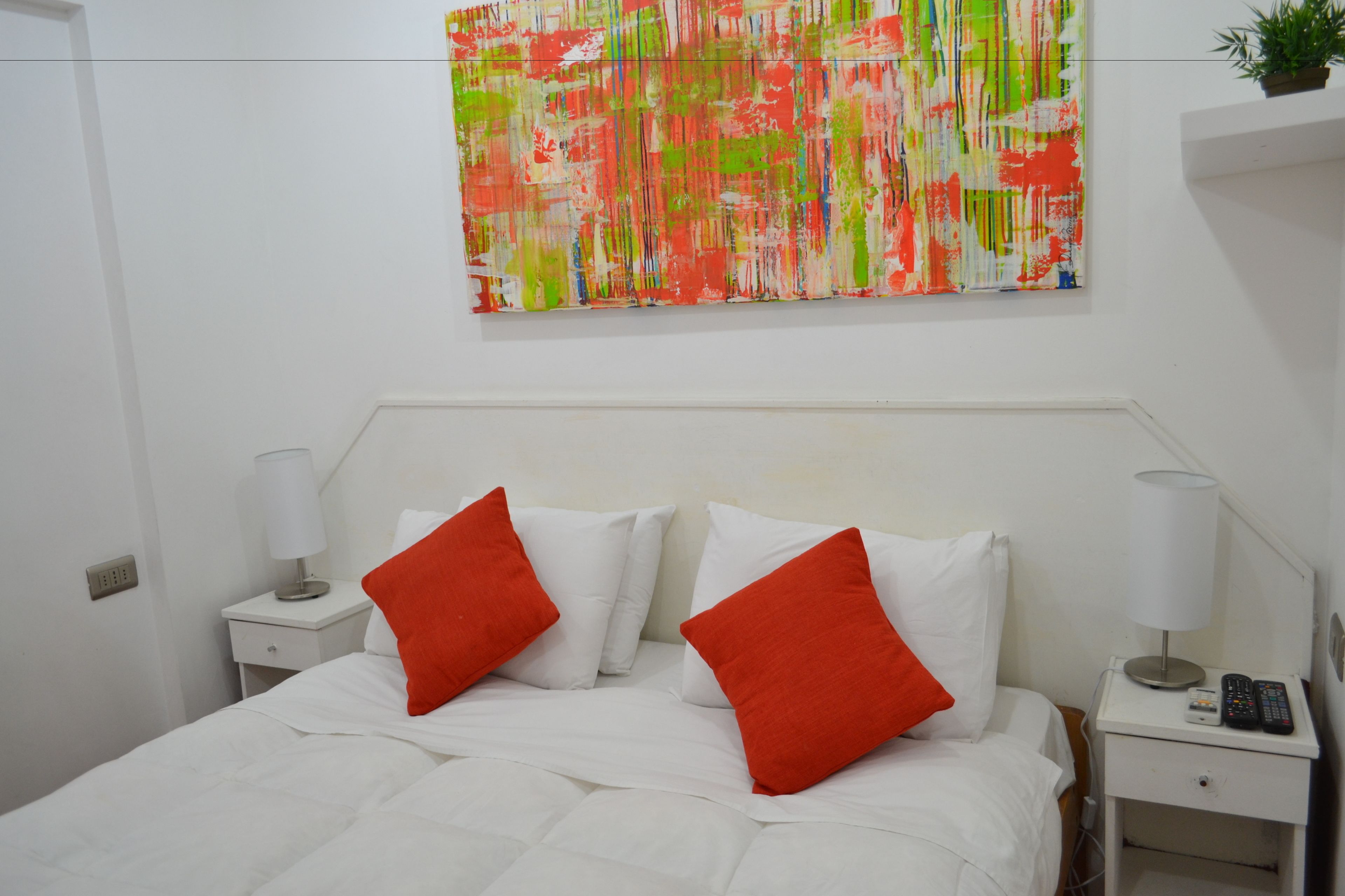 Habitación de hotel con cama, ropa de cama blanca, dos almohadas rojas, cabecero blanco, mesita de noche con lámpara, teléfono y un cuadro abstracto de colores en la pared.