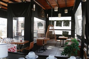 An outdoor dining area with wicker furniture, hanging light fixtures, and large windows with views of greenery. - Casona Bustamante (Santiago)