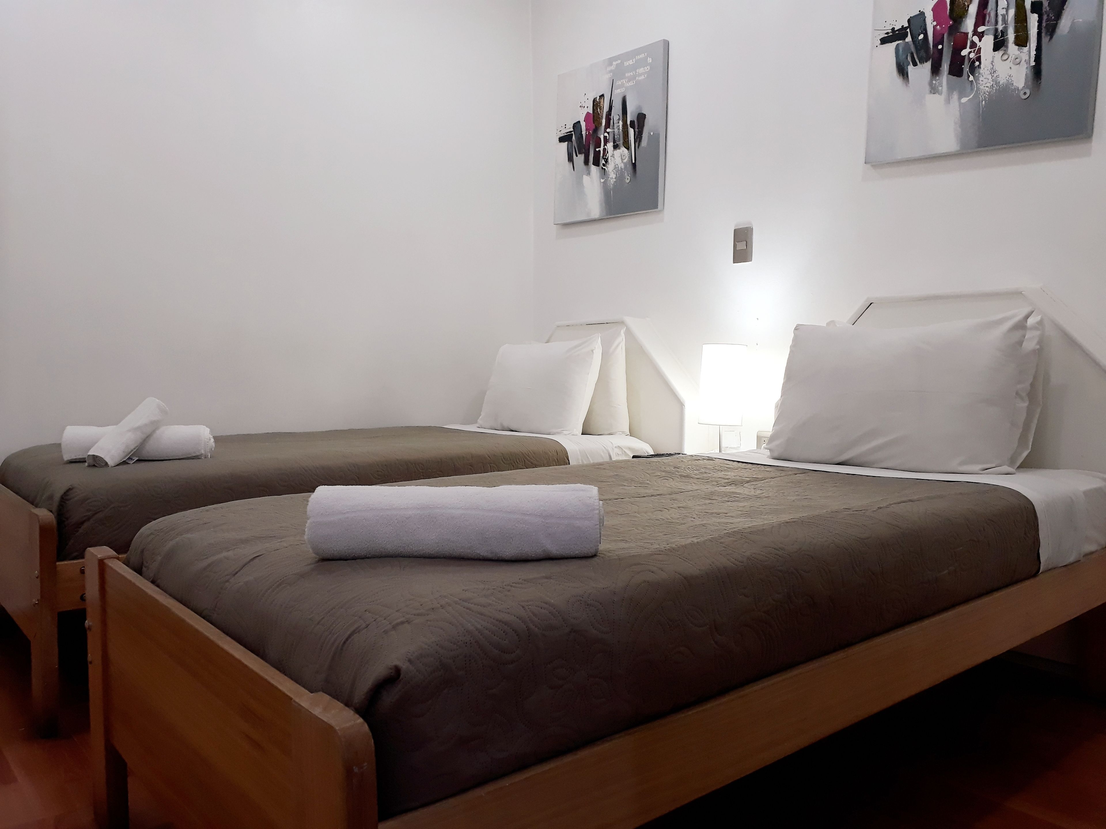 Una habitación de hotel con dos camas individuales, cada una con cubrecama marrón y almohadas blancas. Hay una silla de madera y una mesita con una lámpara.
