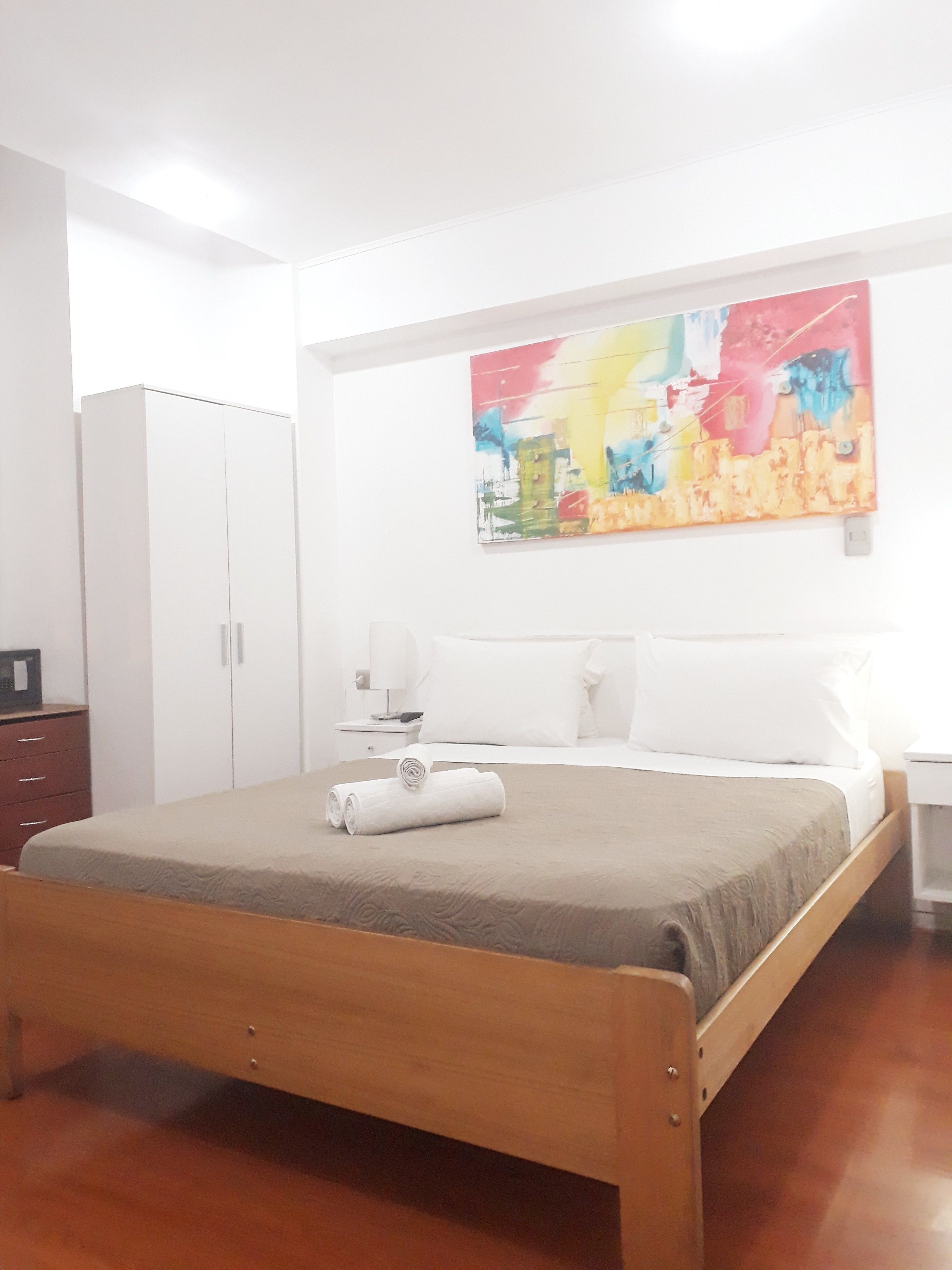 A hotel room with a bed, a wardrobe, a nightstand, a lamp, and a colorful abstract painting on the wall.