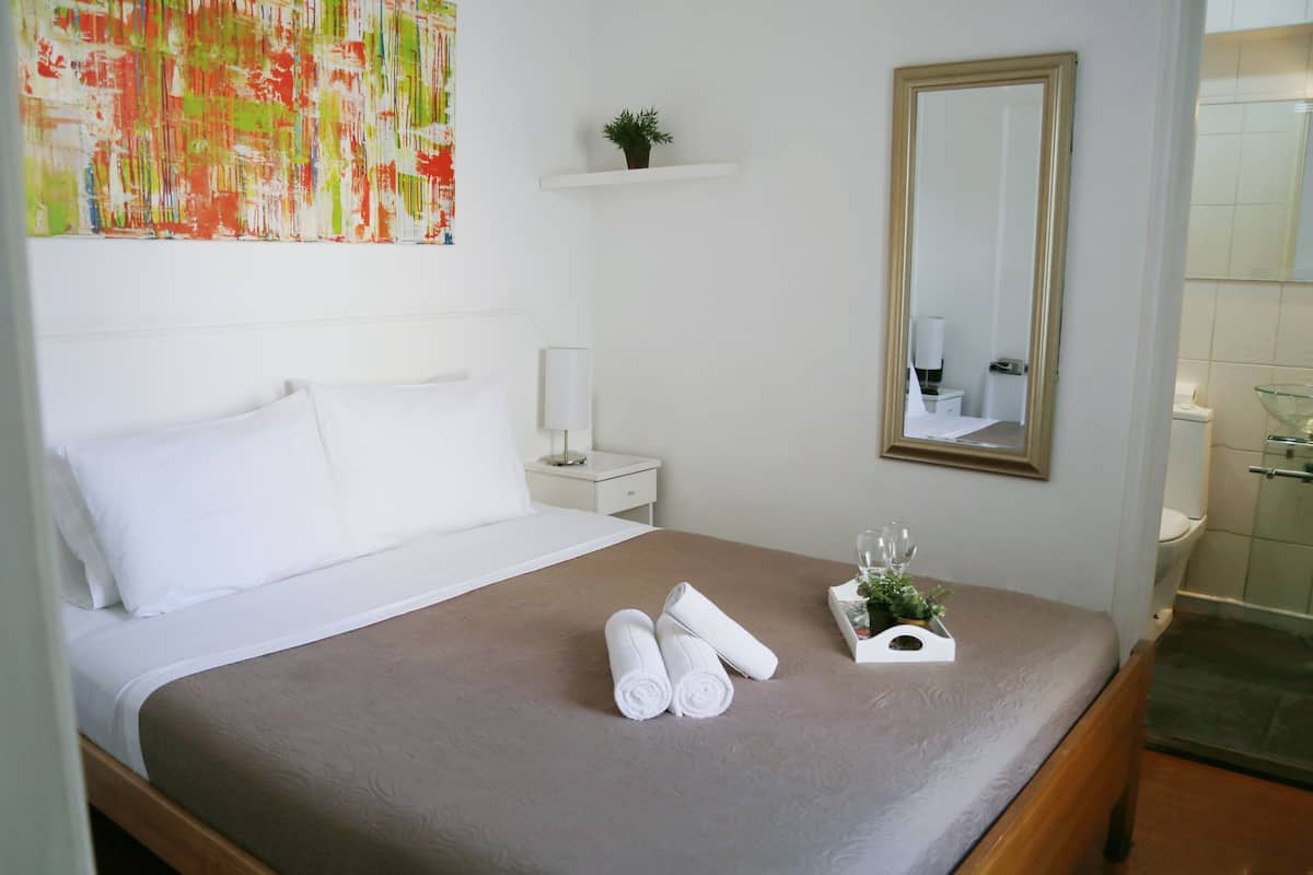 Quarto de hotel com cama, mesa de cabeceira, espelho, prateleira pequena com uma planta e banheiro visível através de uma porta aberta.