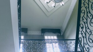 A room with a high ceiling, a decorative ceiling light fixture, a window with a view, and a metal railing with intricate designs. - Hotel Meryland (Colon)