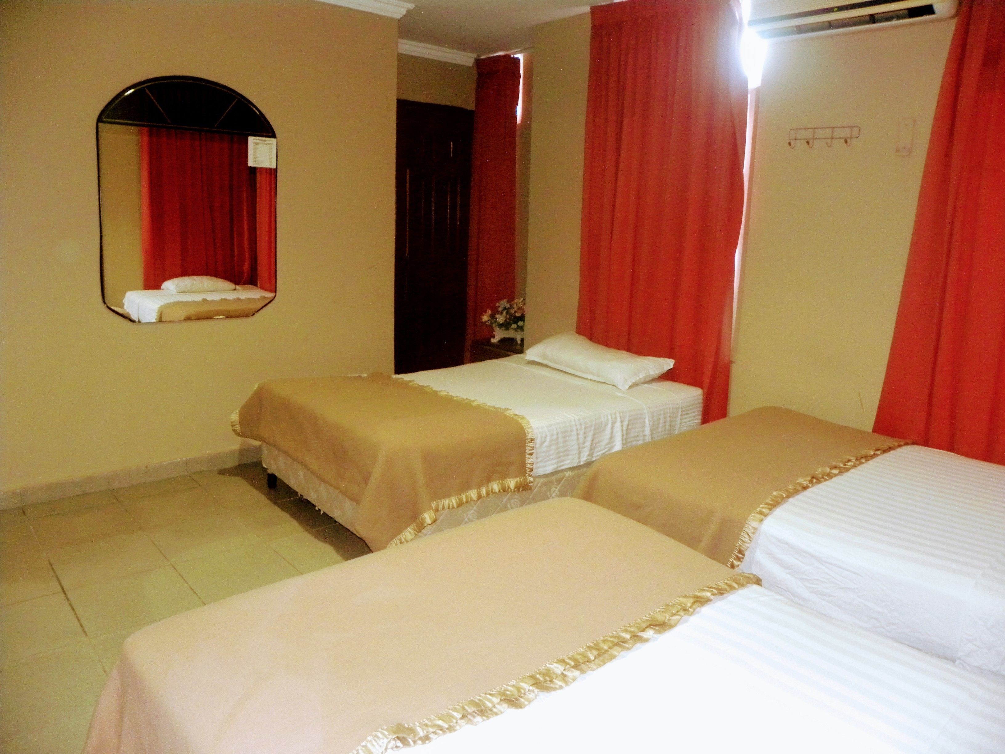 A hotel room with two beds, red curtains, a mirror, and a small table with a flower vase.