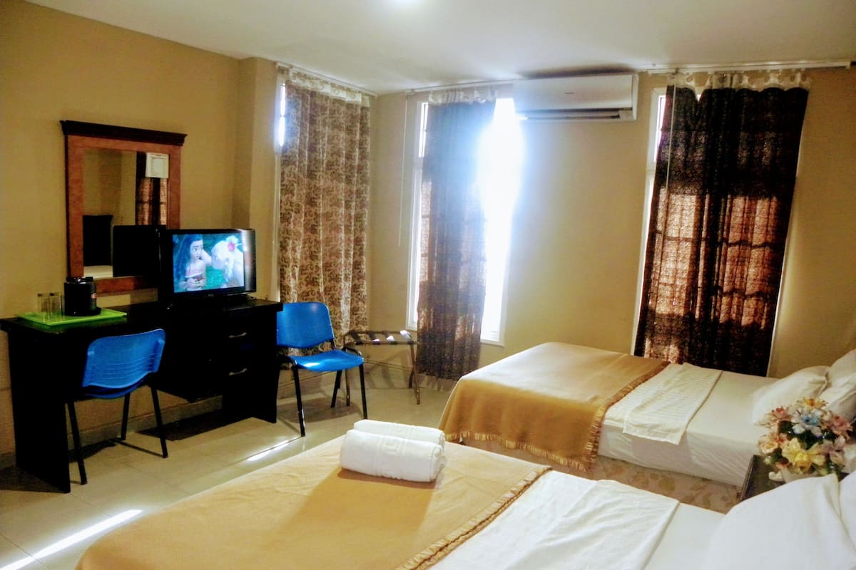 Ein Hotelzimmer mit zwei Betten, einem Schreibtisch mit Fernseher, einem Sessel und einem Fenster mit Vorhängen.