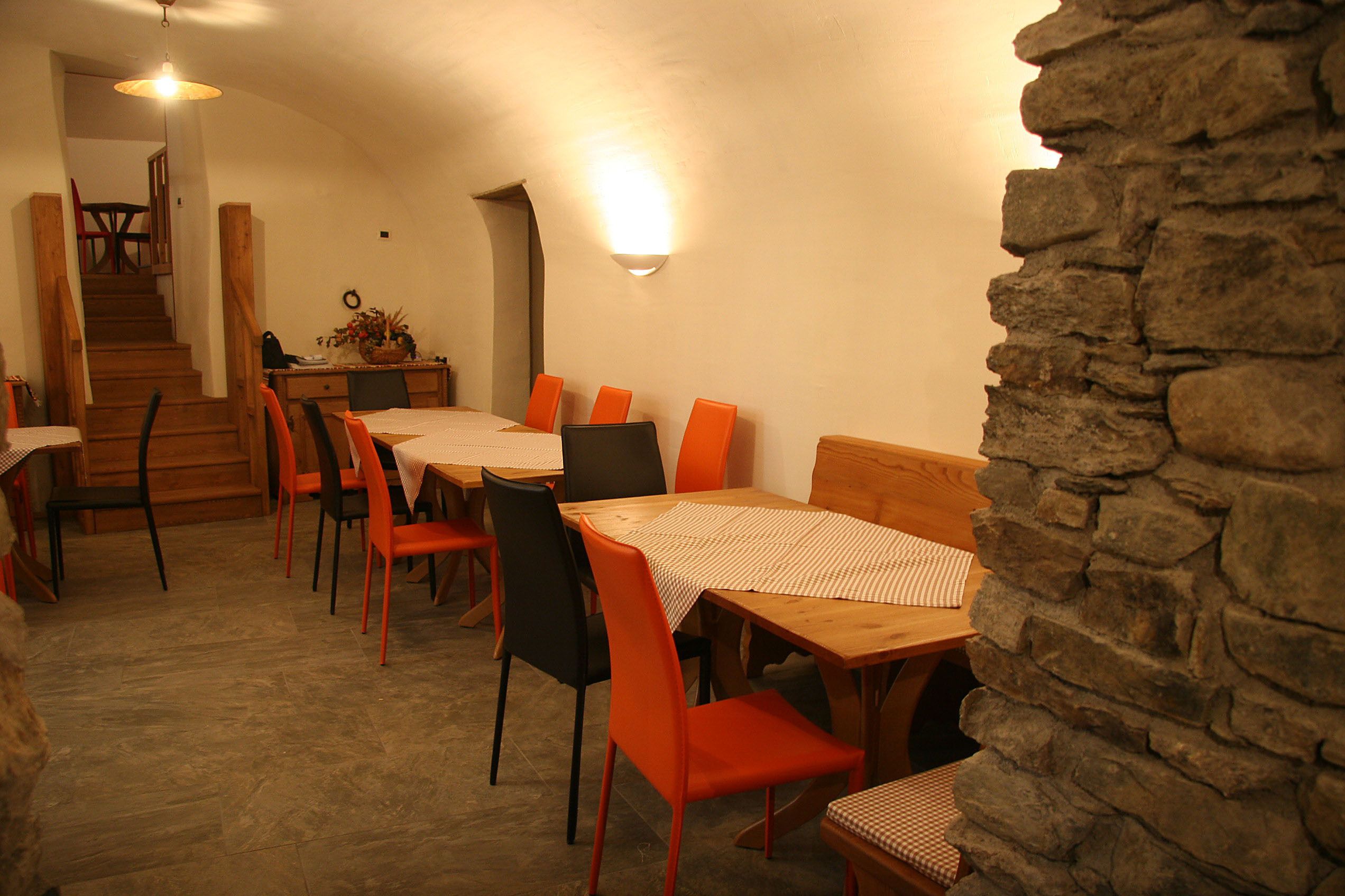 A dining area with wooden tables and chairs, a stone wall, and a staircase leading to another room.