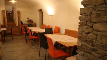 A dining area with wooden tables and chairs, a stone wall, and a staircase leading to another room.