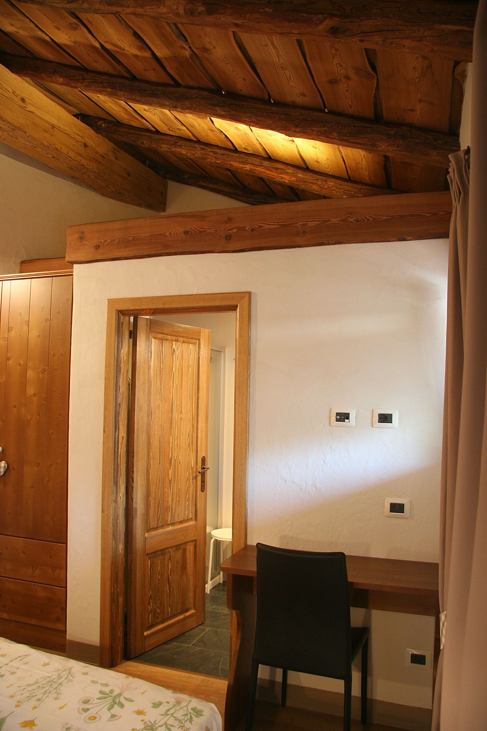 A room with a wooden ceiling, a wooden door, a desk with a chair, and a bed with a floral bedspread.