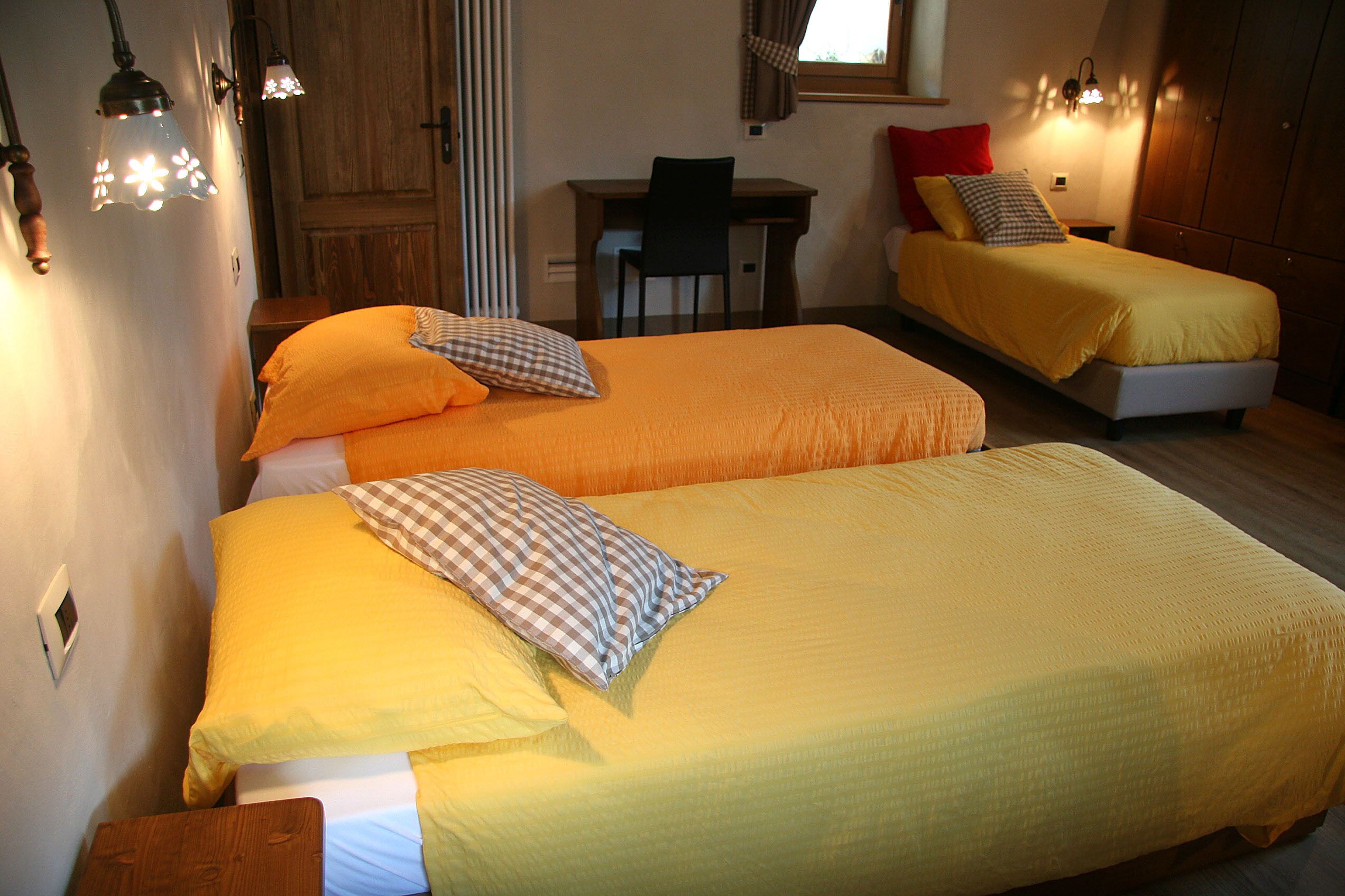 A room with two beds, each with yellow bedding and checkered pillows. There is a desk with a chair and a window with curtains.