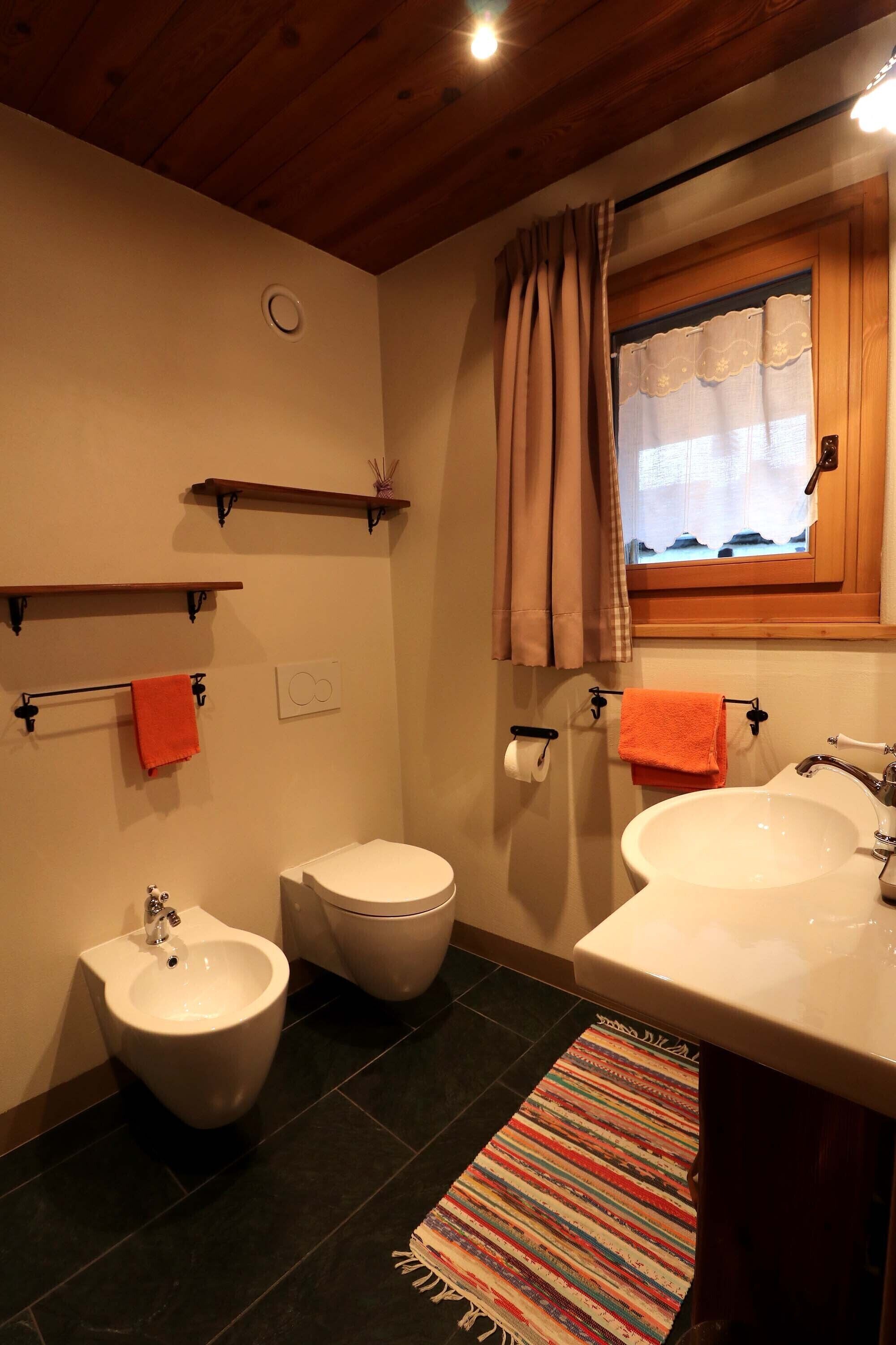 Ein Badezimmer mit integriertem Bidet, Toilette und Waschbecken, Holzregalen, einem Fenster mit Vorhängen und einem farbenfrohen Teppich.