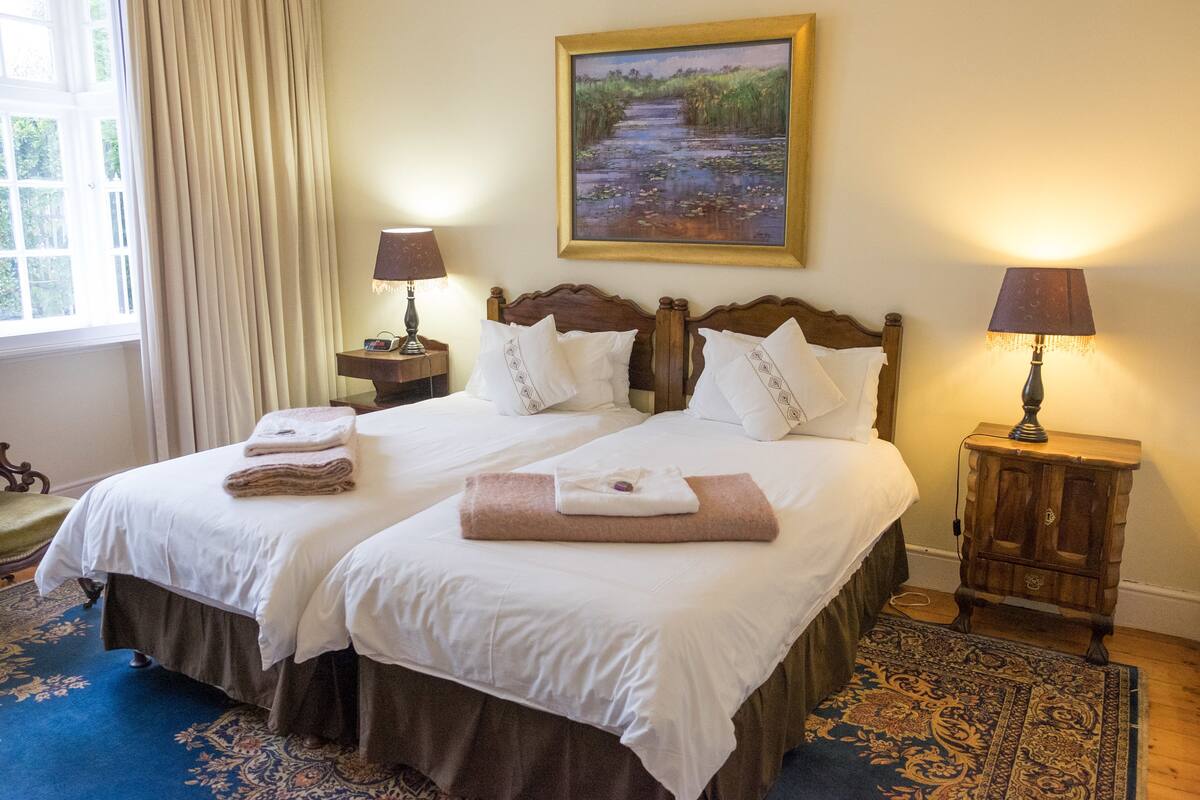 A double bed with white linens, two bedside tables with lamps, a painting of a landscape, and a blue patterned rug.