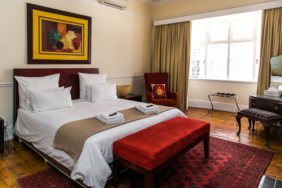 A neatly made bed with white linens and a red bench, a wooden chair with a patterned cushion, and a large window with curtains.