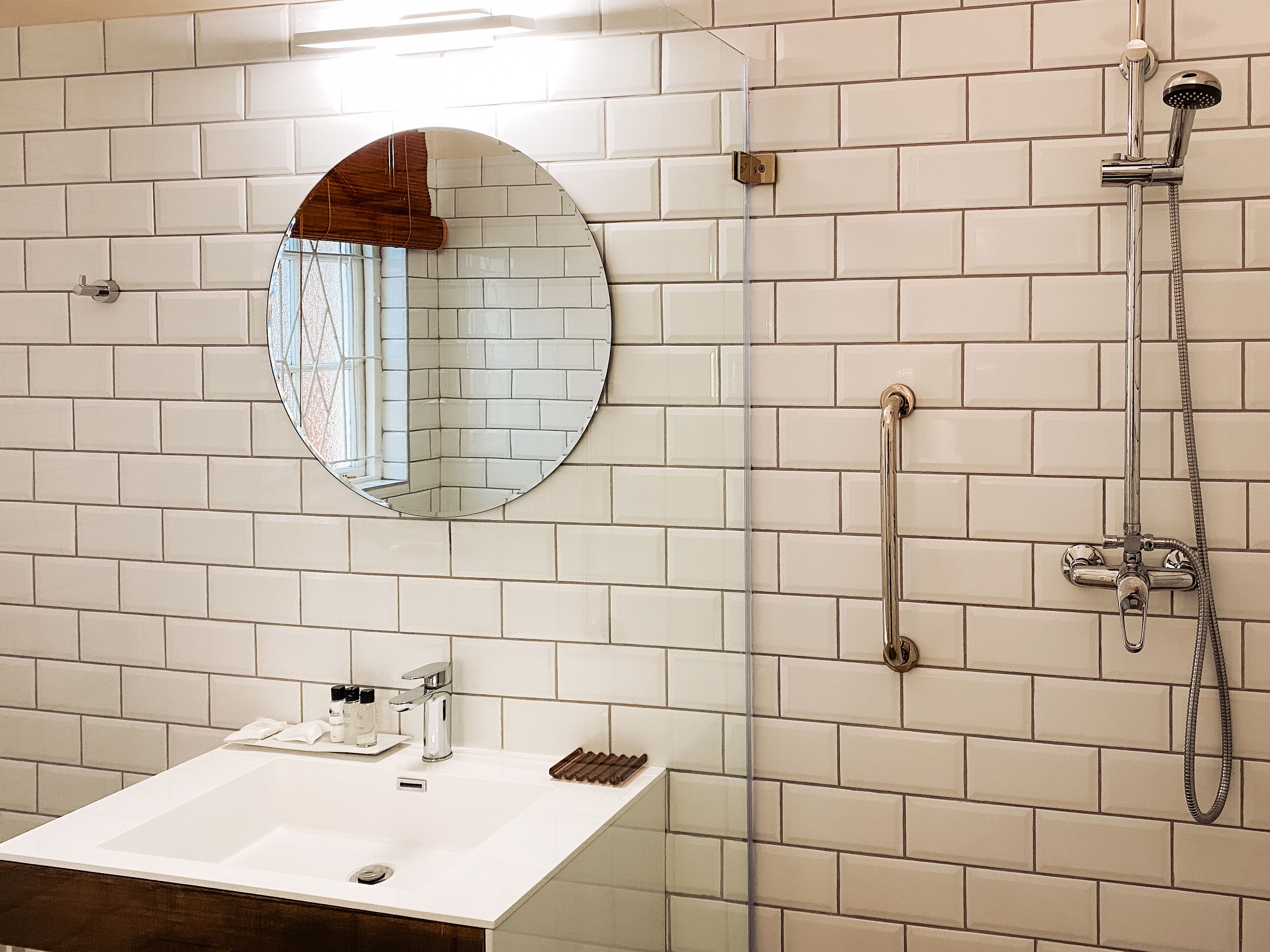 Un baño con paredes de azulejos blancos, un espejo redondo, un lavamanos con artículos de aseo y una ducha con cabezal de ducha manual.