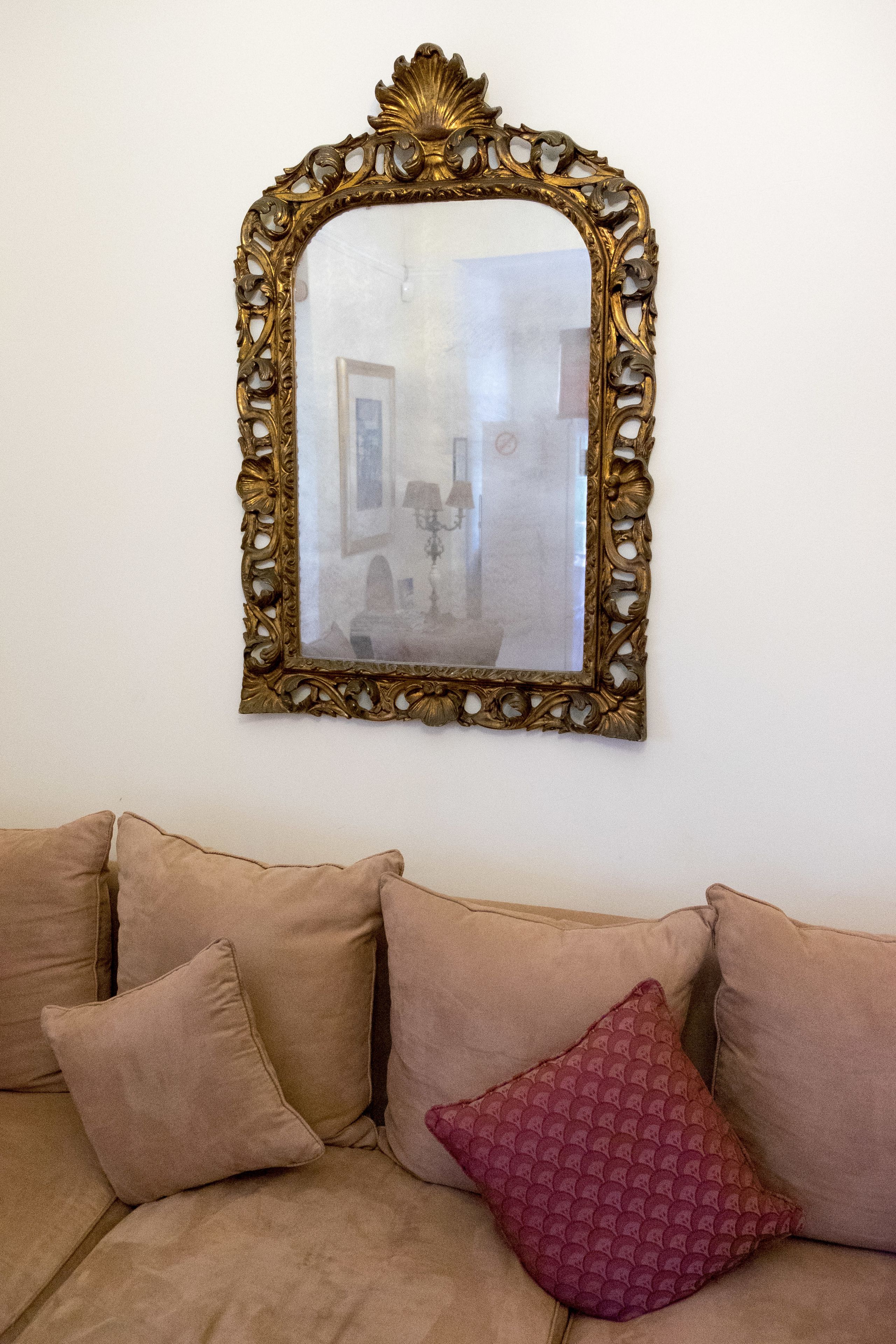 Una habitación con un sofá color crema, un espejo decorativo con un marco dorado ornamentado y un cojín estampado.