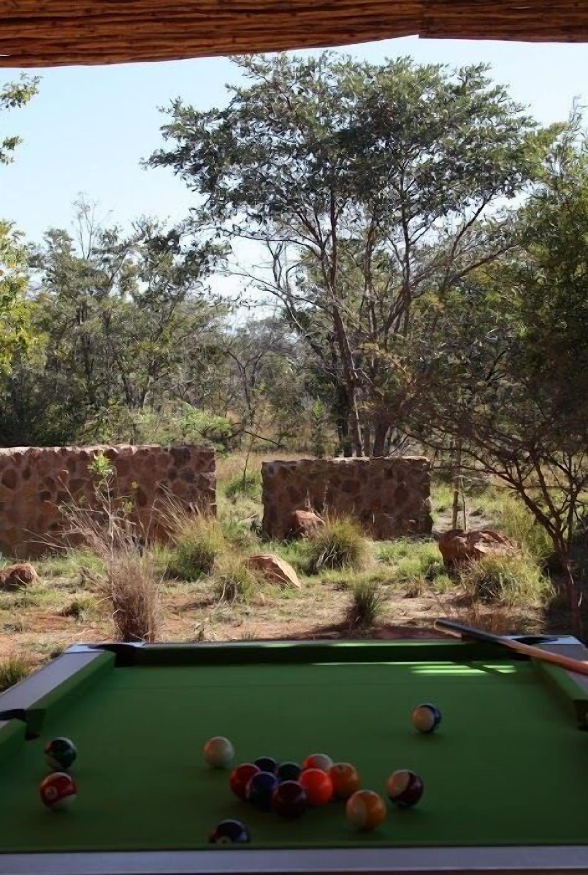 A pool table with balls, set against a natural backdrop with trees and stone walls.