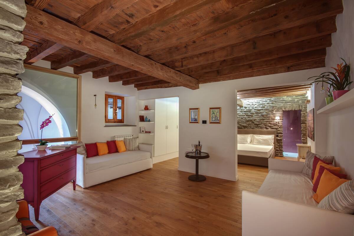 Ein gemütliches Wohnzimmer mit Holzbalken, einer Steinwand und einem weißen Sofa mit bunten Kissen.