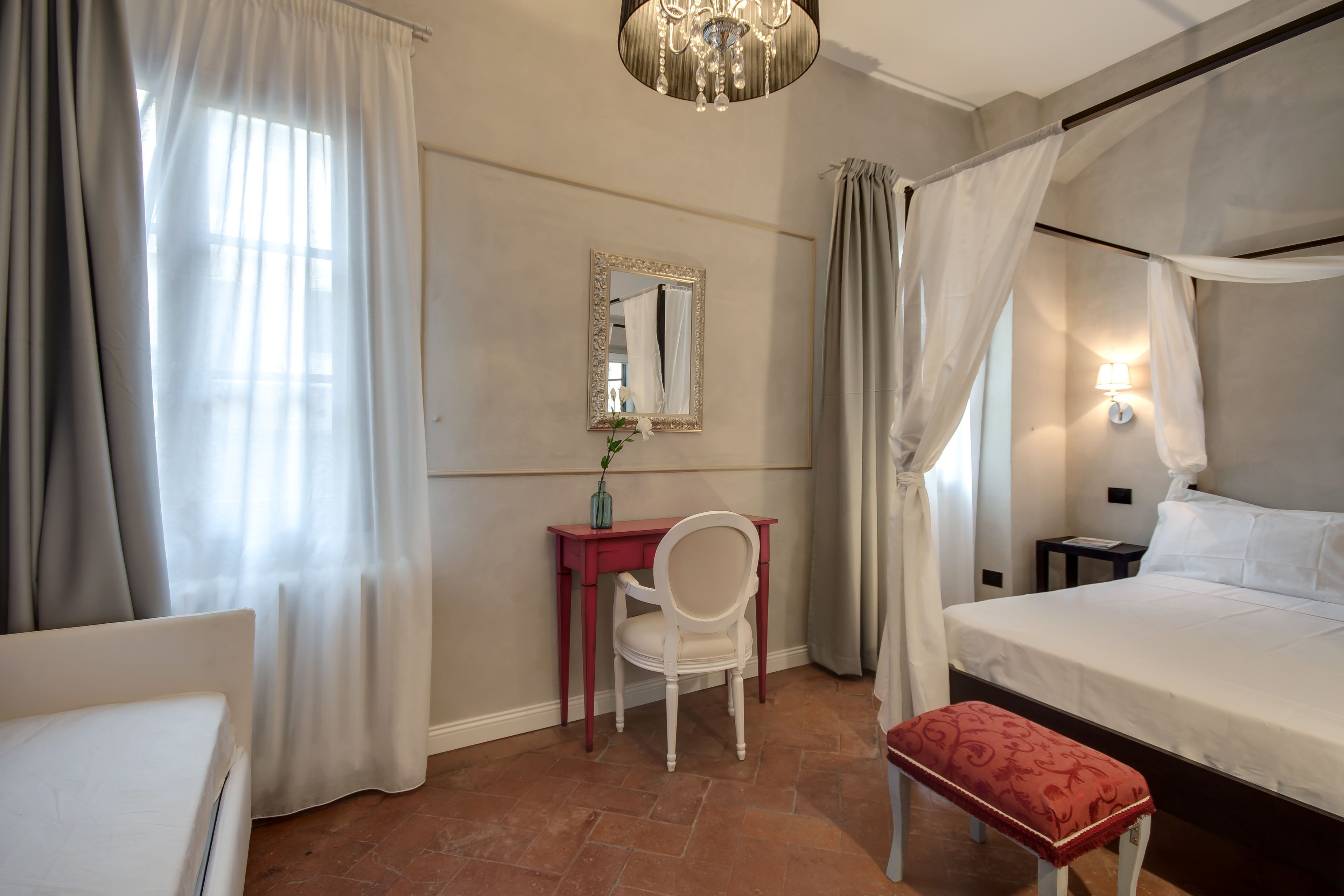 A hotel room with a bed, a desk with a chair, a bedside table, a nightstand, a red bench, and a chandelier.