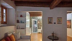 A cozy living room with a wooden ceiling, a white sofa with colorful cushions, a small round table with a glass, and a kitchen visible through an open doorway.