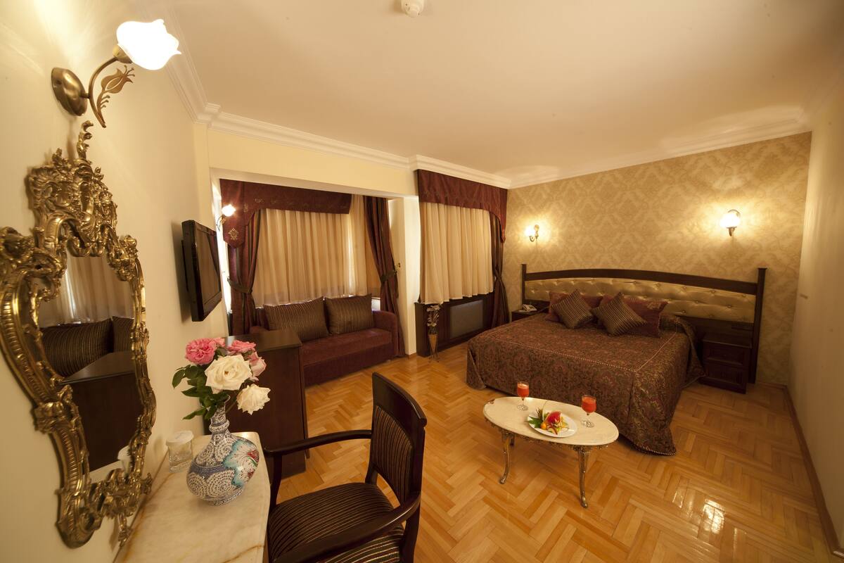A hotel room with a bed, a sofa, a dining table, a chair, a vase with flowers, a mirror, and a television.