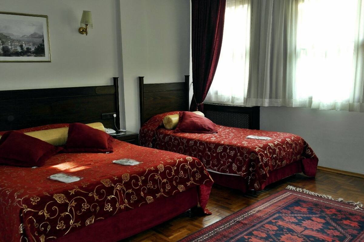 A hotel room with two beds, a large window with curtains, a framed picture on the wall, and a patterned rug on the floor.
