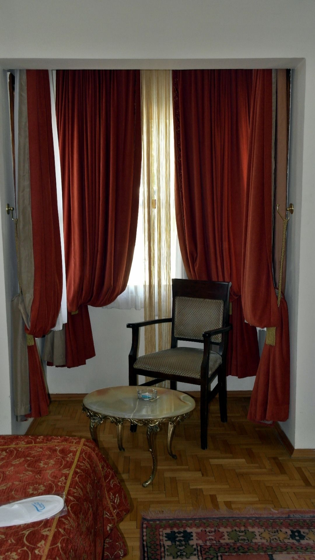 A room with a wooden floor, a round table with a glass top, a chair with a woven seat, and a bed with a red patterned cover.