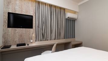 A hotel room with a flat-screen TV mounted on a wooden wall, a desk with a remote control, and an air conditioning unit.