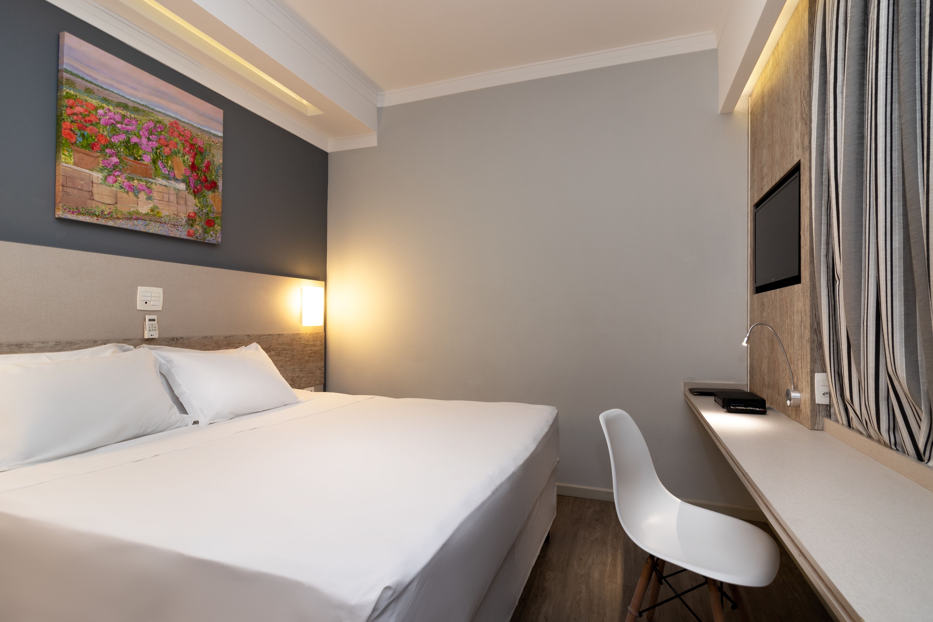 A hotel room with a bed, bedside table, desk, chair, and a painting on the wall.