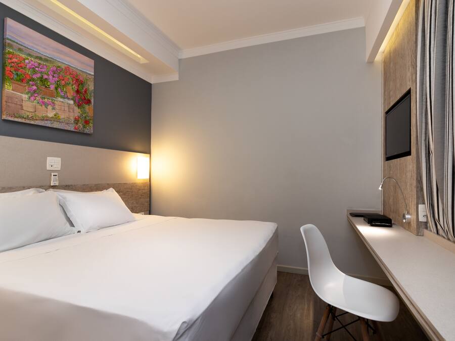 A hotel room with a bed, bedside table, desk, chair, and a painting on the wall.