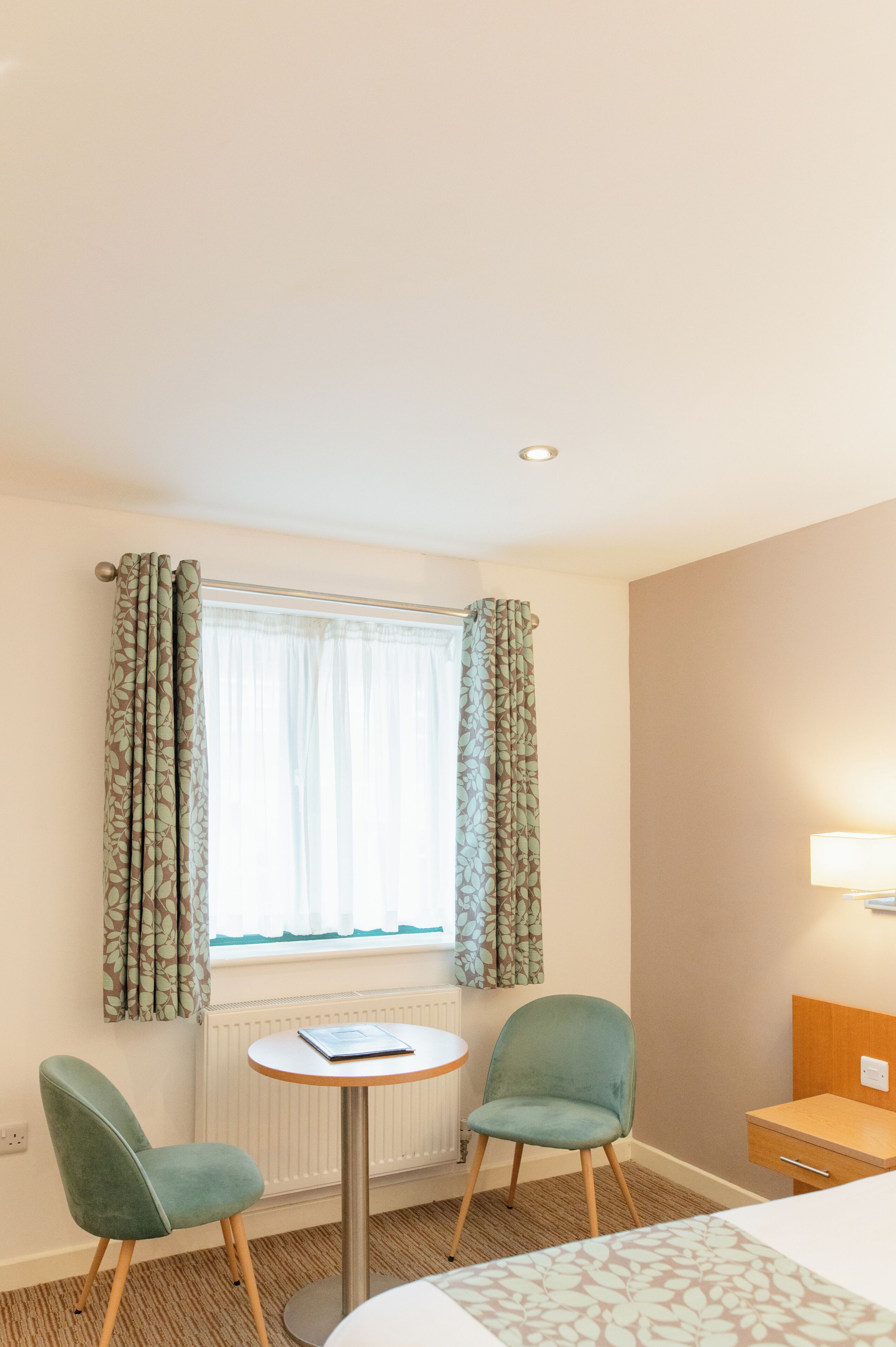 A hotel room with a bed, a small round table, two chairs, a window with curtains, a radiator, and a bedside lamp.