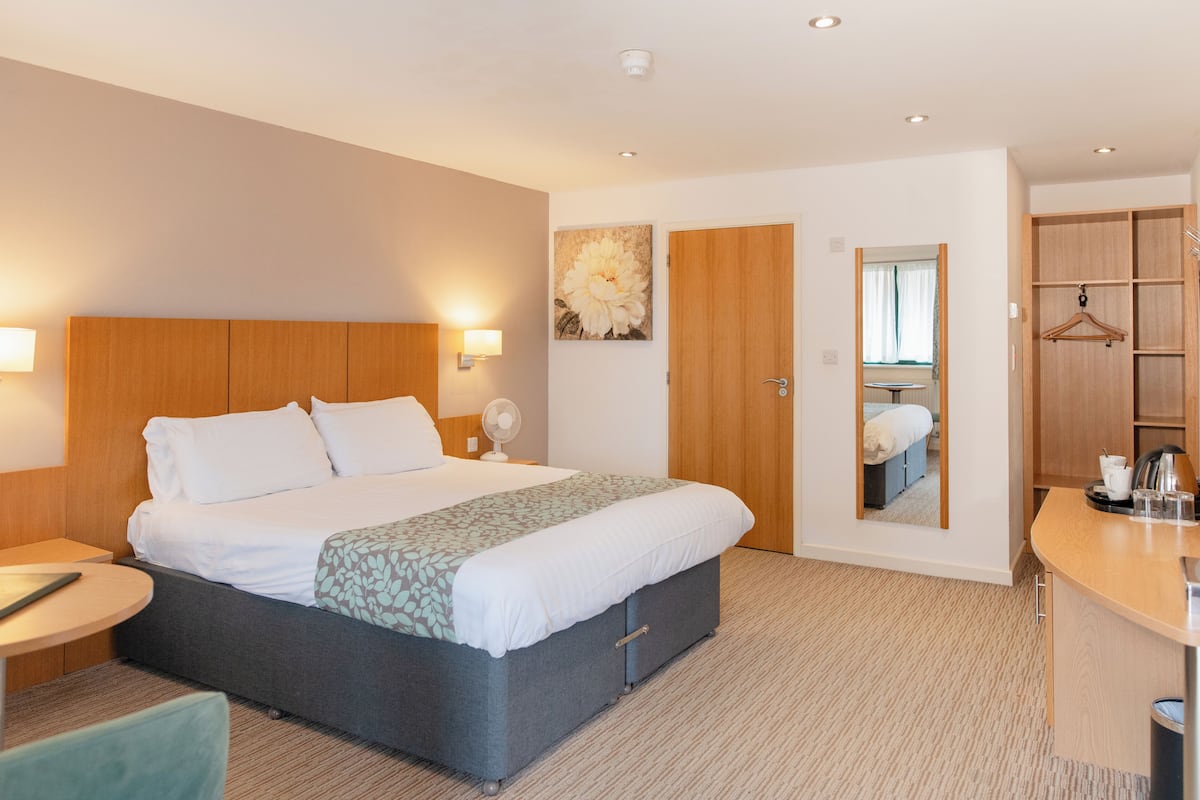 A hotel room with a large bed, bedside table, lamp, and a mirror. There is a wooden wardrobe and a small table with a kettle and cups.