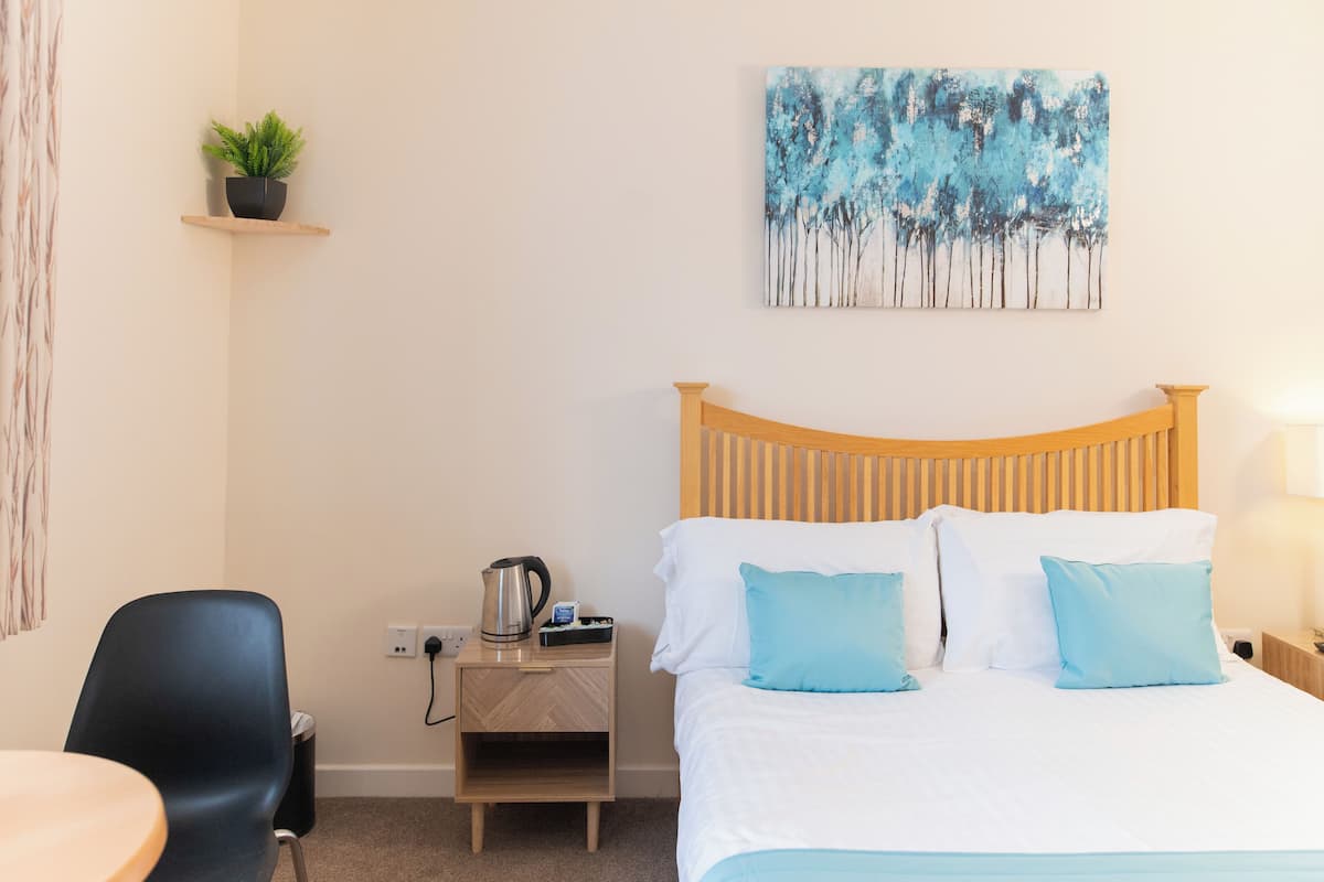 A hotel room with a wooden headboard, a bed with white and blue bedding, a nightstand with a kettle, a small potted plant, and a painting of trees on the wall.