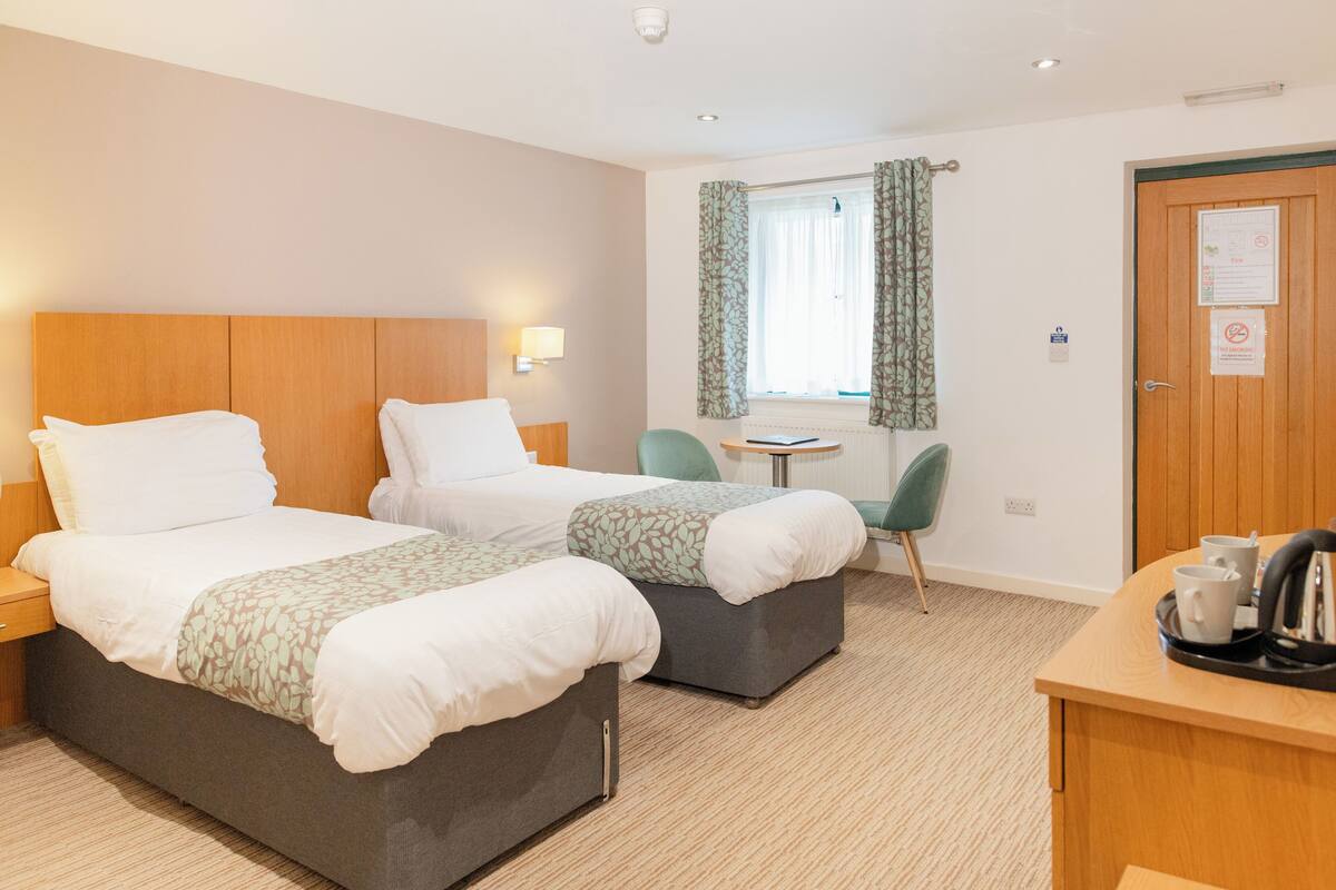 A hotel room with two beds, a small table with chairs, a window with curtains, and a door with a sign.