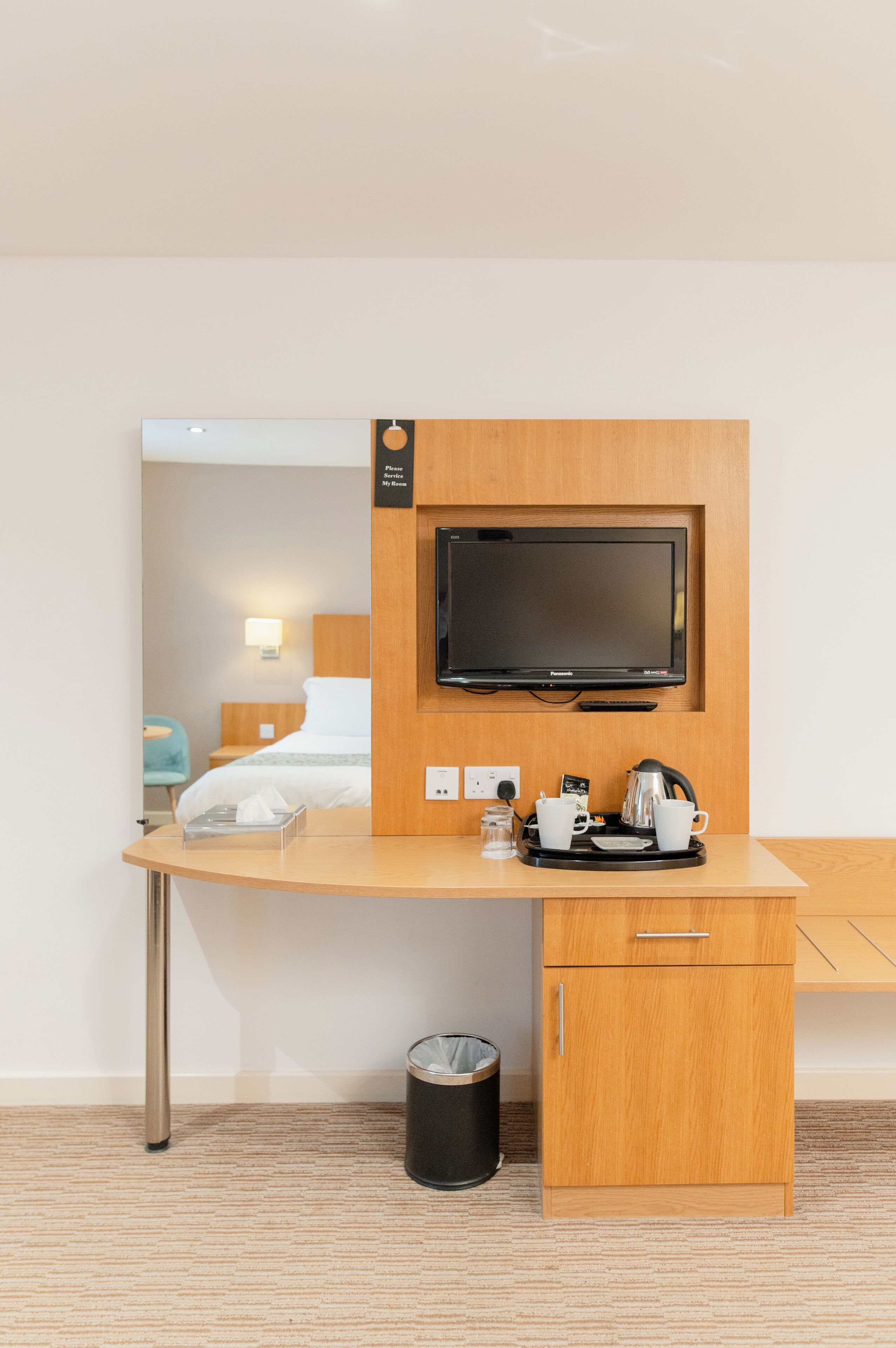Una habitación de hotel con un escritorio de madera, un televisor, una cama, un bote de basura y un armario pequeño.