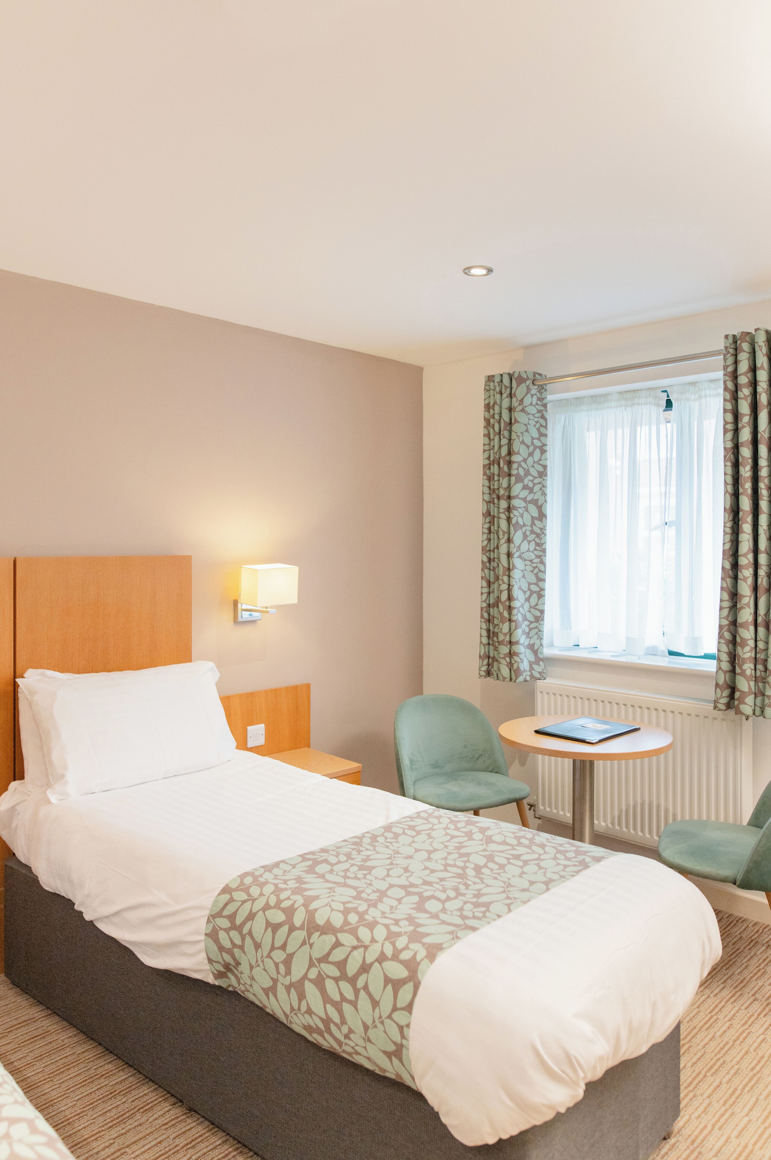 A hotel room with a bed, a nightstand, a lamp, a chair, a small table, and a window with curtains.