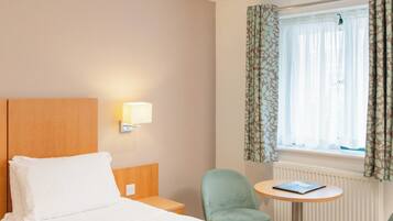 A hotel room with a bed, a nightstand, a lamp, a chair, a small table, and a window with curtains.