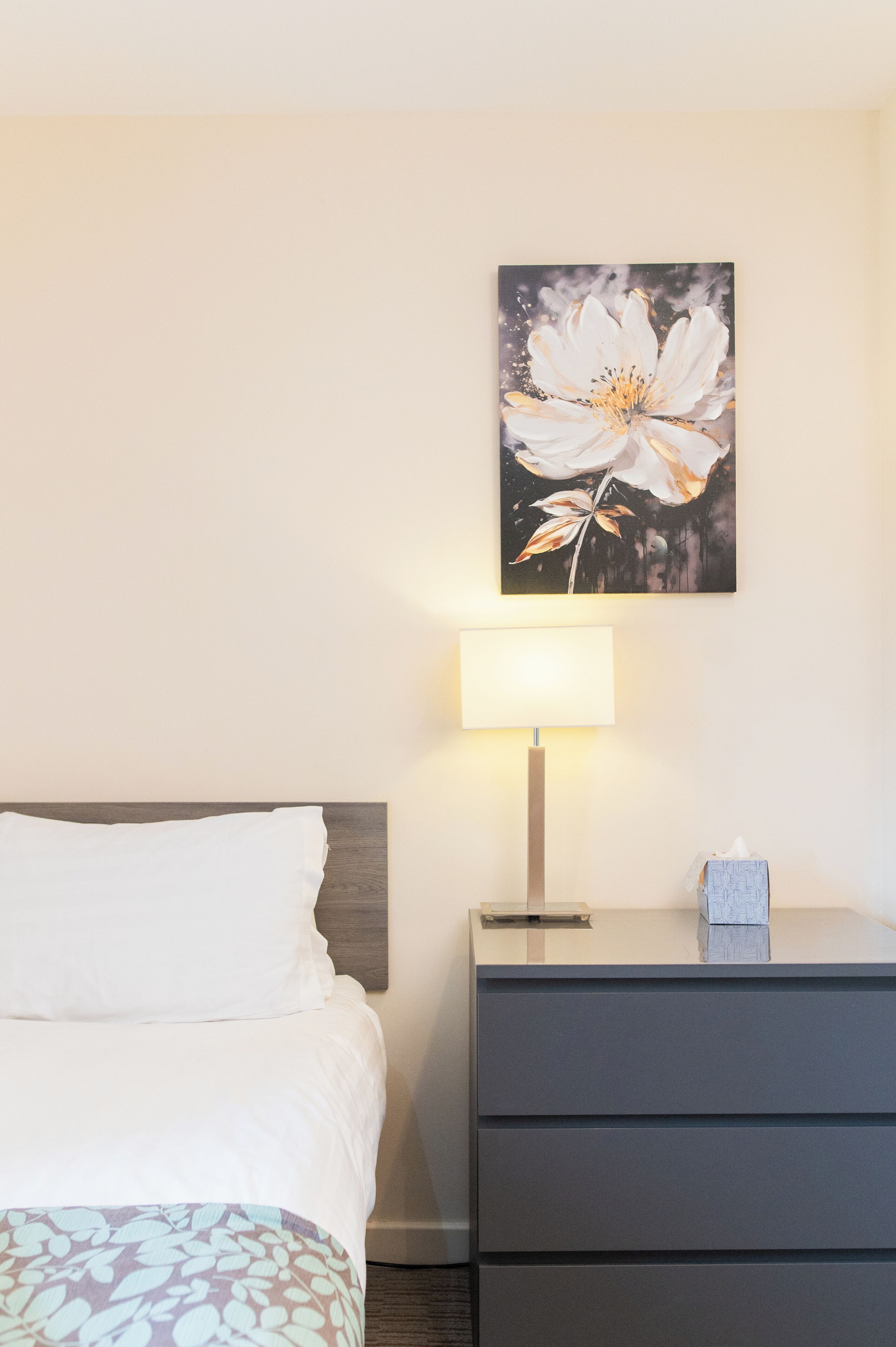 Une chambre d’hôtel avec un lit, une table de chevet avec une lampe, une commode et une œuvre d’art représentant une fleur, accrochée au mur.
