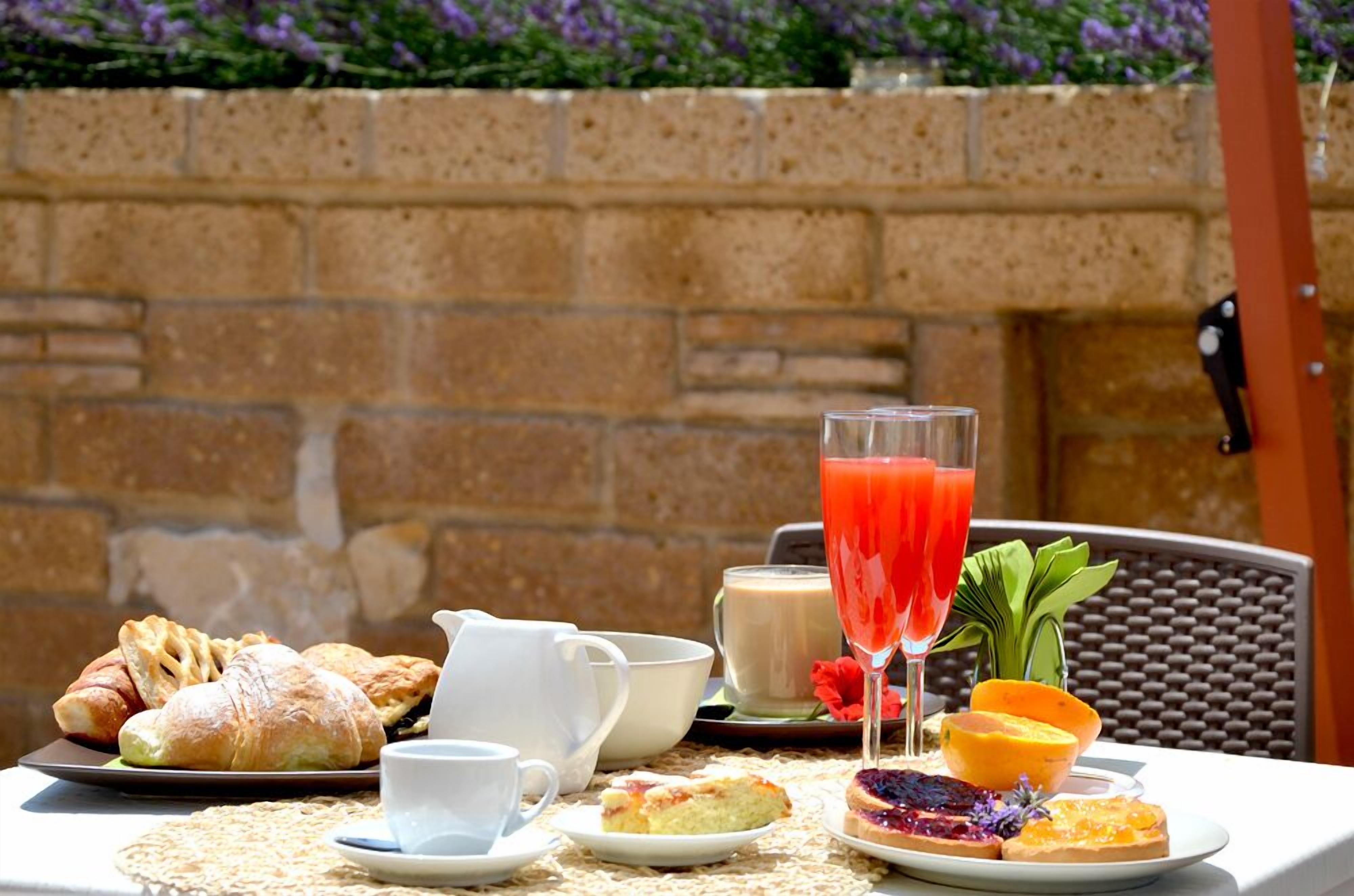 Une table dressée avec des pâtisseries, une carafe, des tasses et un verre de jus, avec un mur de briques et des plantes en arrière-plan.