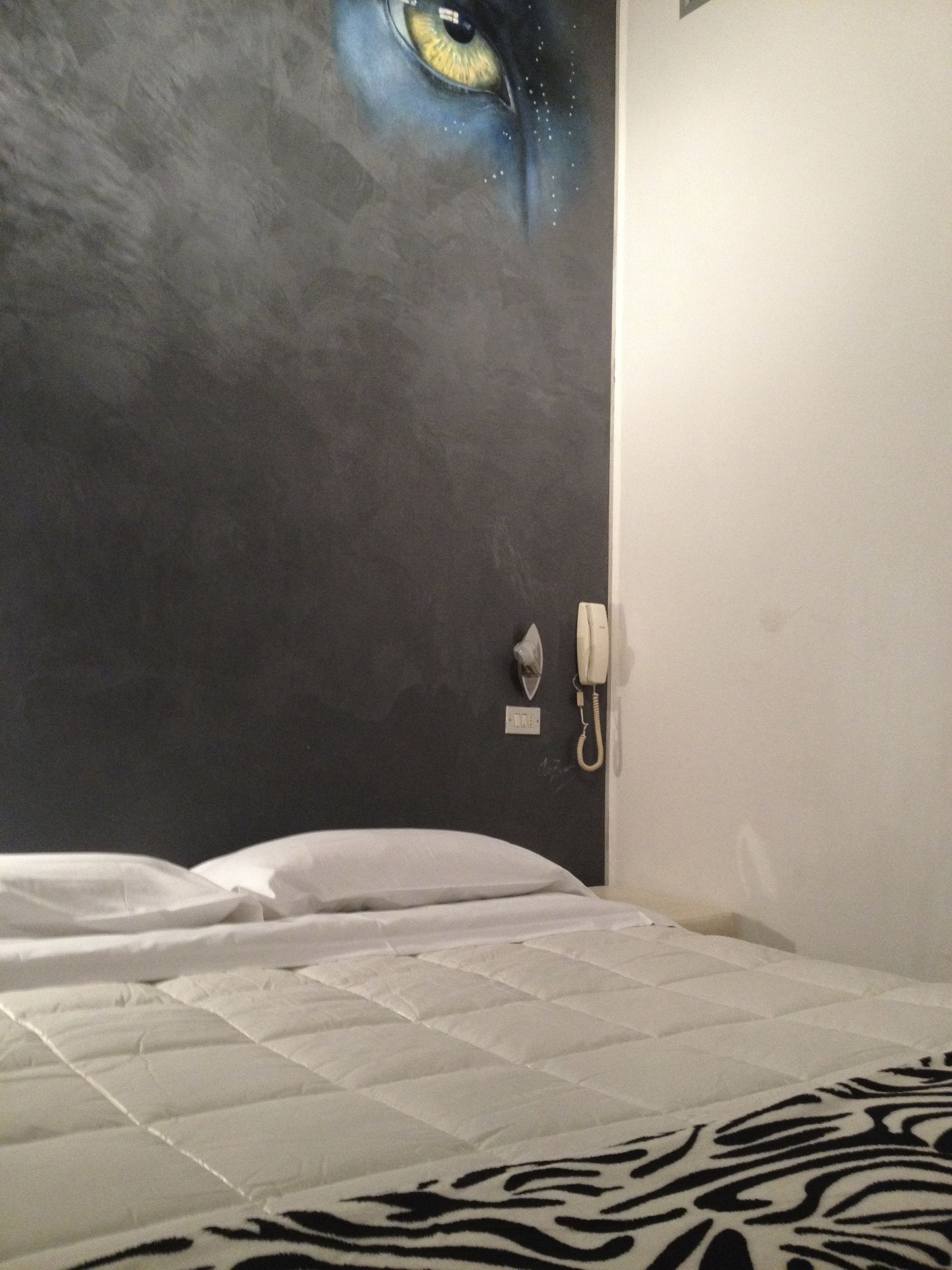 Ein Hotelzimmer mit einem Bett, einer Zebra-Decke, einer Wand mit einem Augen-Gemälde, einem Telefon und einem Lichtschalter.