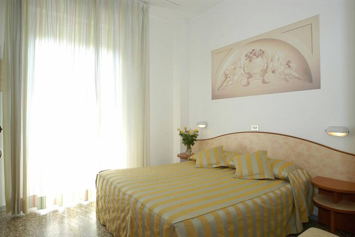 Une chambre d’hôtel avec un lit, une table de chevet, une lampe fixée au mur et une fenêtre avec des rideaux.
