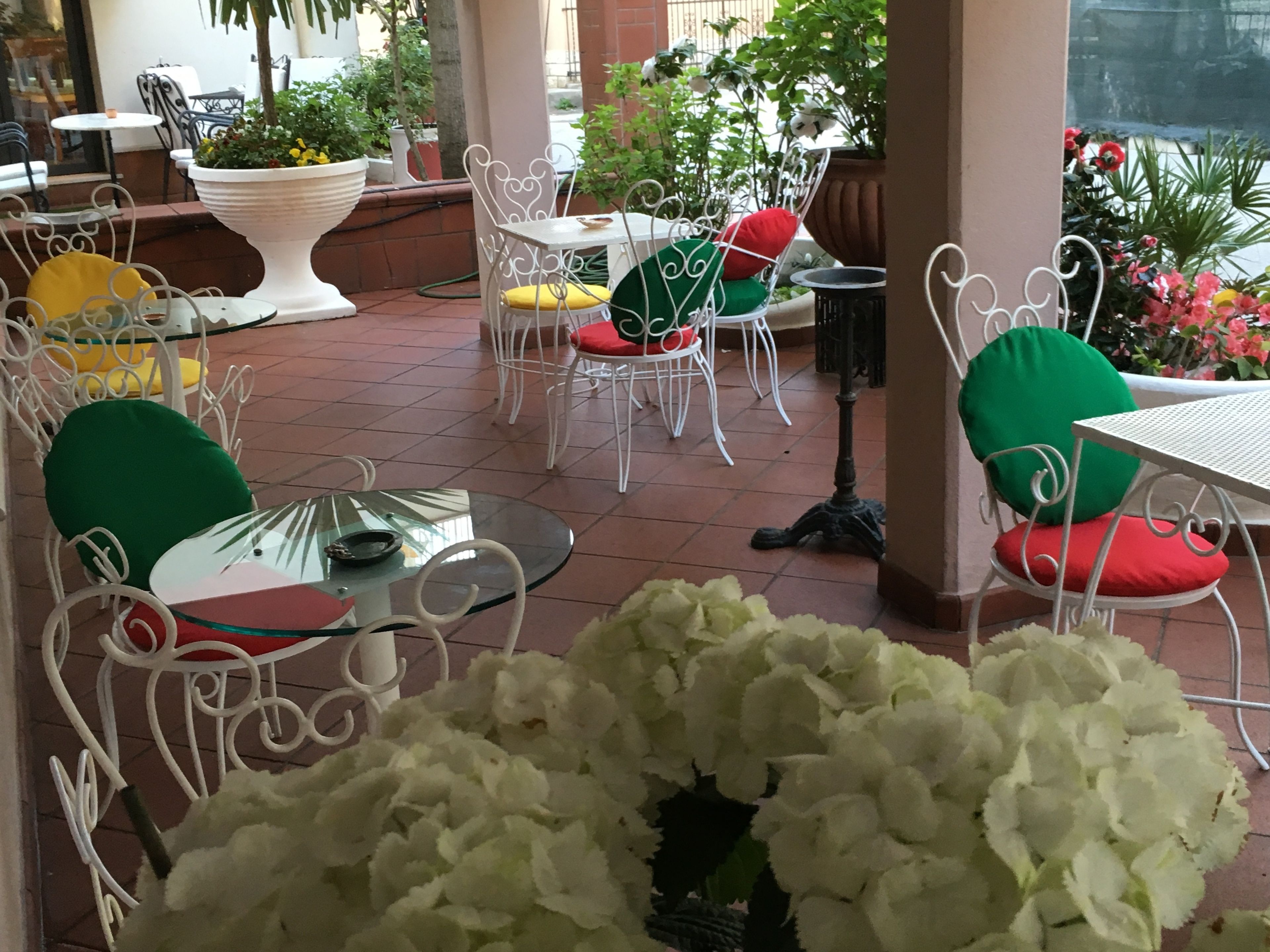 An outdoor patio with white metal chairs and tables, surrounded by potted plants and flowers.